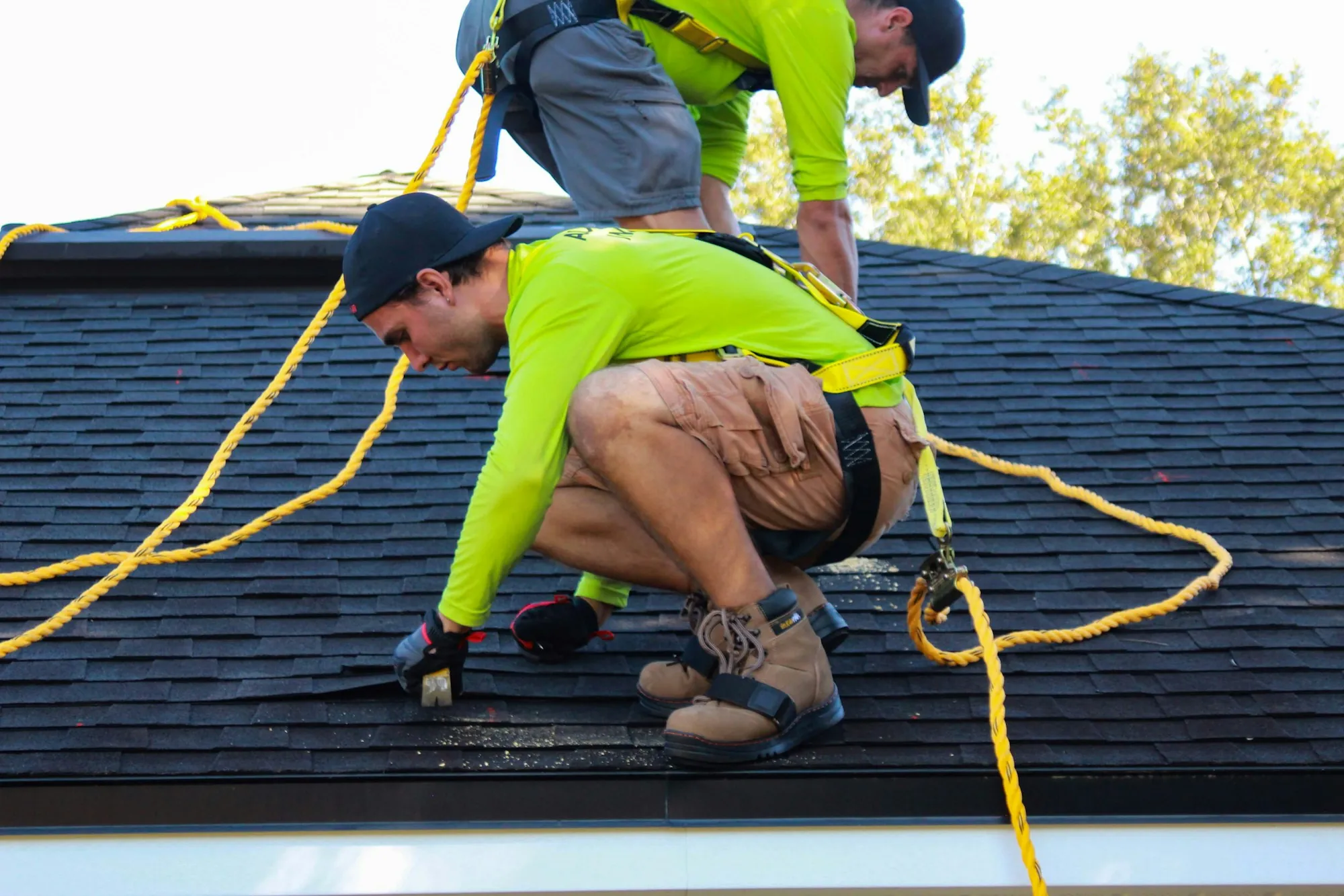 Roofer in a safety harness kneeling to work on an asphalt shingle roof